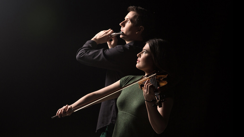 Photo of woman in green with a violin and man in black with a flute, against a dark background and looking into a bright light.