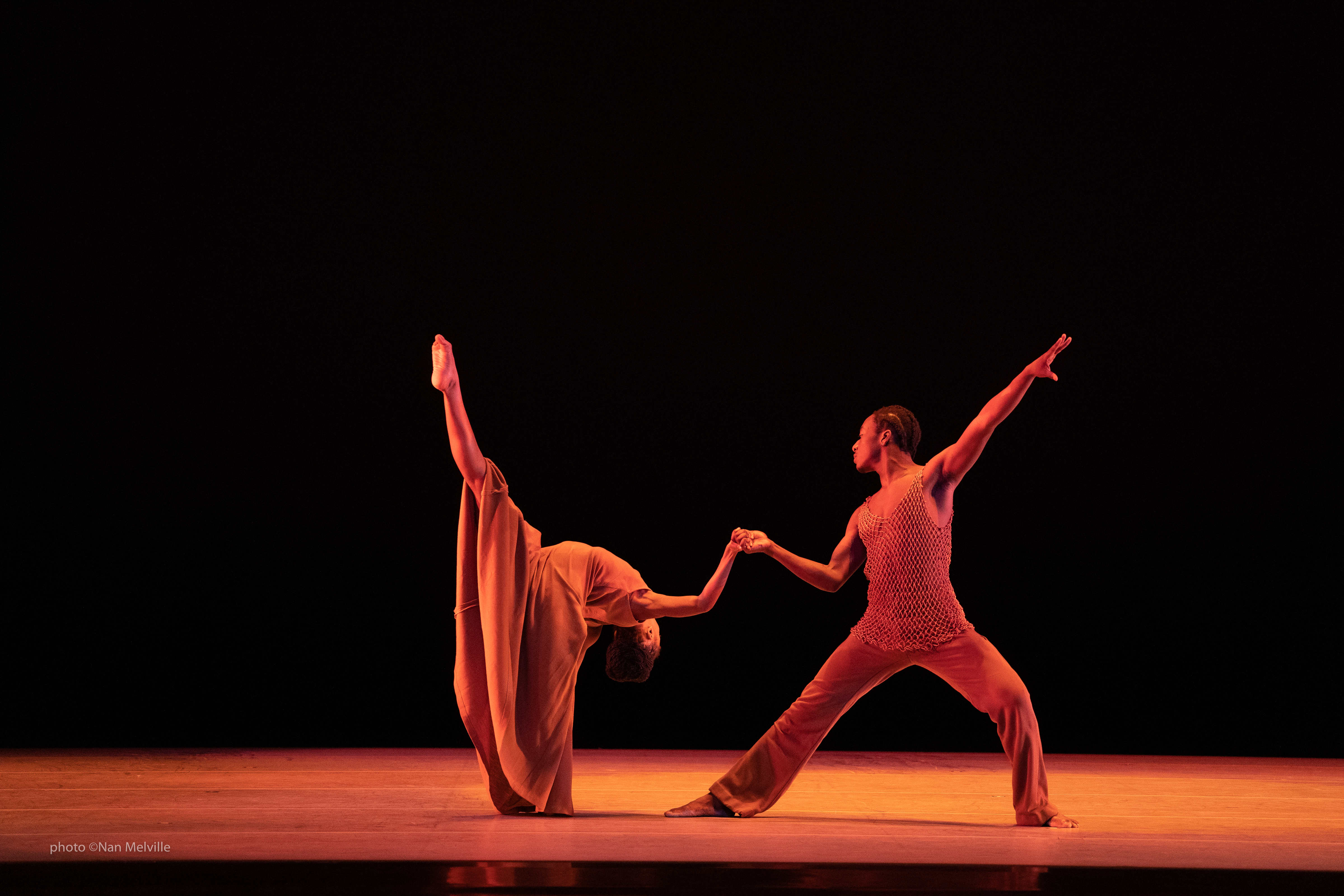 Photo of two dancers posing on stage under an orange light, against a black background.